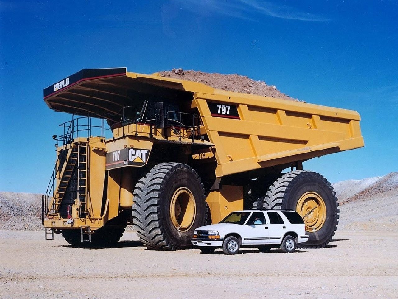 A large Caterpillar dump truck towers over a small SUV, both parked in a barren landscape, highlighting the scale of heavy machinery in contrast to everyday vehicles.