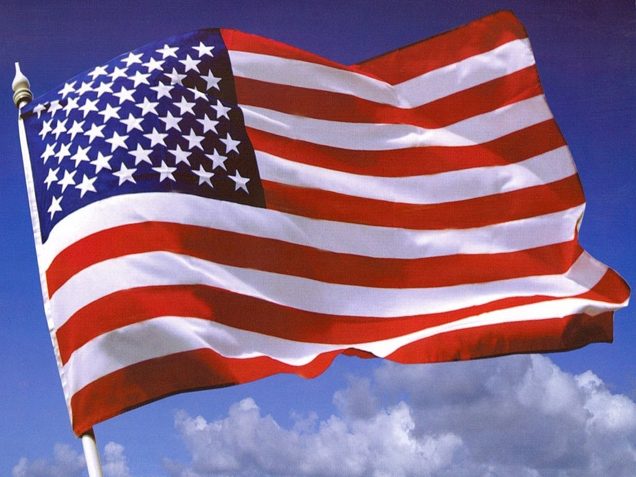 Bright American flag made of fabric waves against a clear blue sky with scattered clouds.