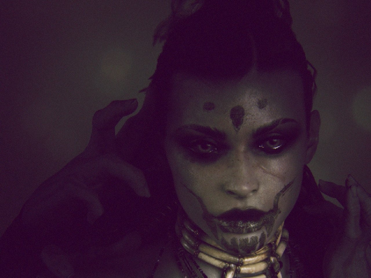 Dark woman with ritual face paint and hollowed eyes, fingers raised near her head, wearing layered bone-like necklaces and bathed in dim, moody greenish light.
