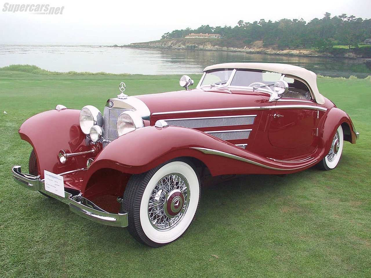 Maroon Mercedes-Benz 500K roadster with whitewall tires, chrome grille and tan soft top, parked on grass beside a coastal shoreline.