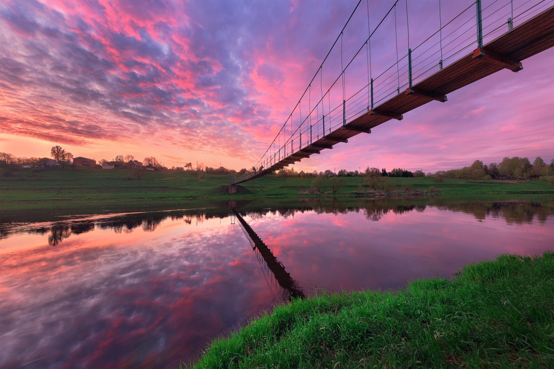 A man-made suspension bridge stretches over a calm river at sunset; vivid pink and purple sky reflects in the water, with grassy riverbank and rolling landscape beyond.