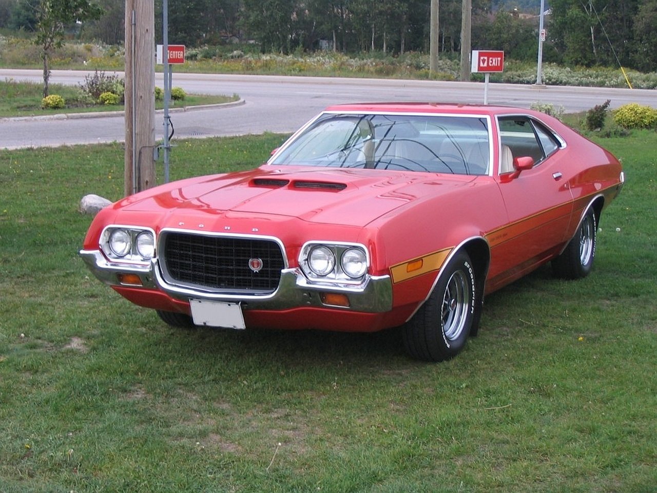 A bright red Ford Gran Torino Sport parked on grass near a roadside, showcasing its classic muscle car design and distinctive front grille.