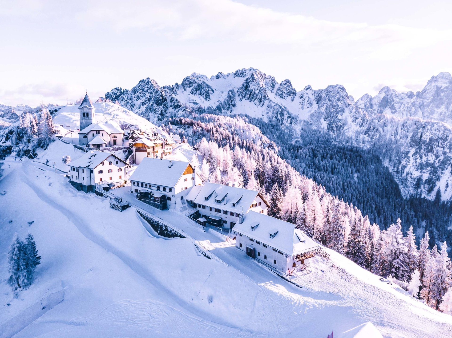 A snowy mountain village with man-made houses nestled on a hillside surrounded by tall, rugged peaks and snow-covered trees.