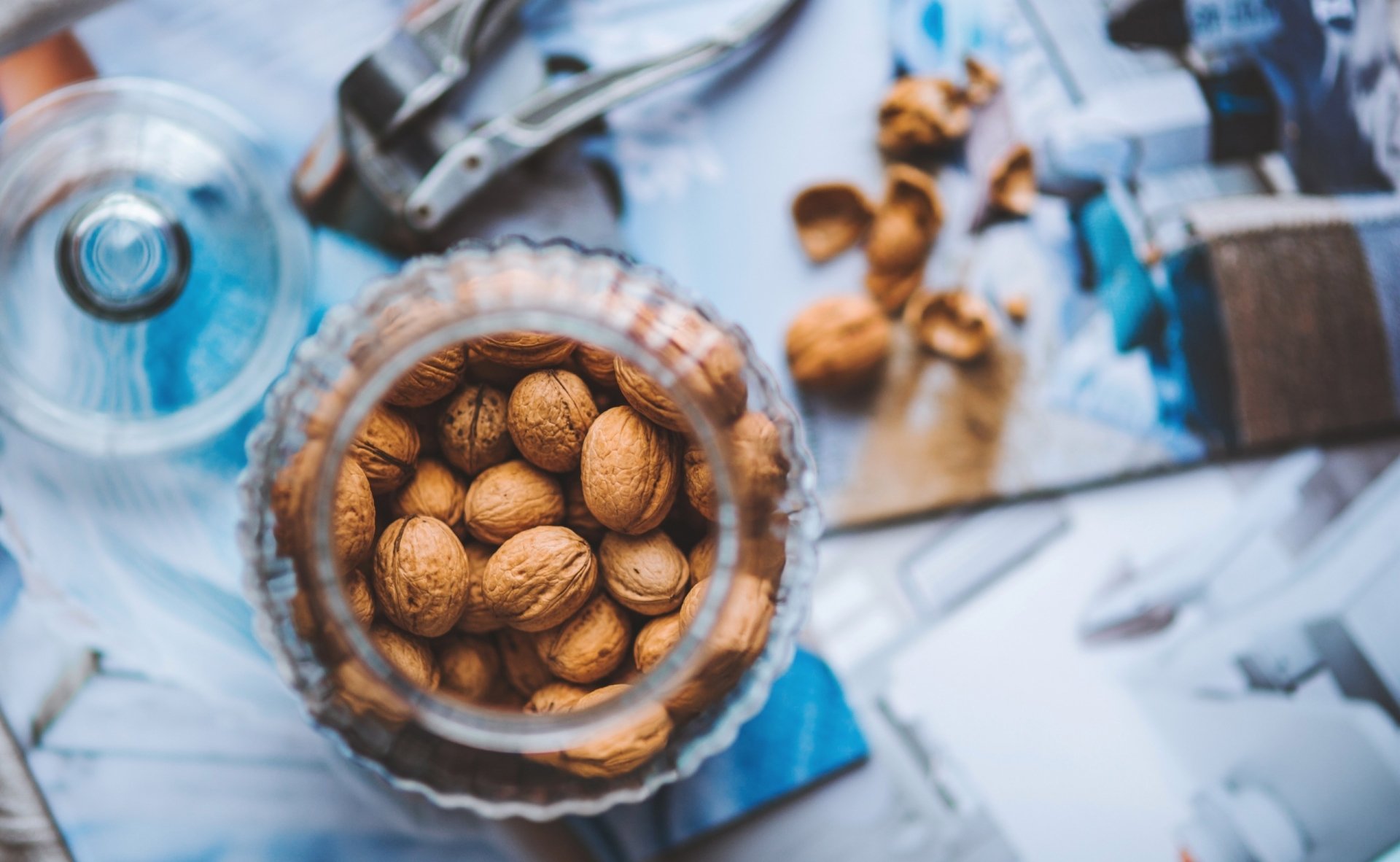 Glass jar of whole walnuts on a tabletop scattered with cracked shells and a nutcracker, a walnut nut food preparation scene.