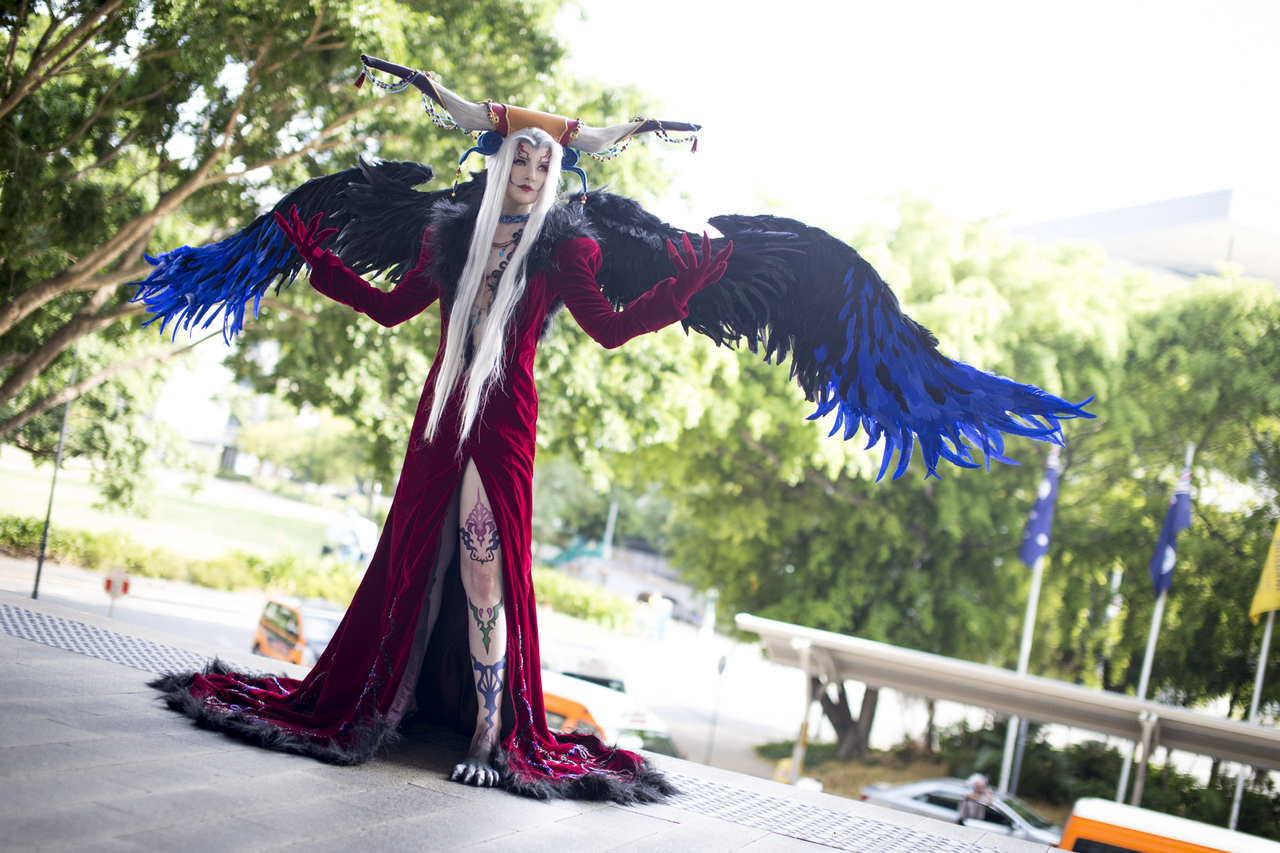 Woman in cosplay wearing a long red dress and horned headpiece, long white hair and dramatic black-and-blue feathered wings outstretched, posing outdoors on steps.