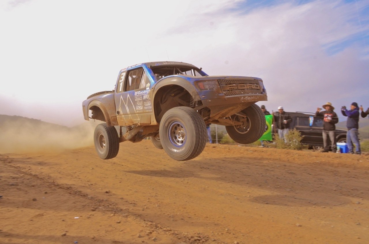 A Ford Raptor vehicle airborne while racing on a dirt track, kicking up dust with several spectators in the background.