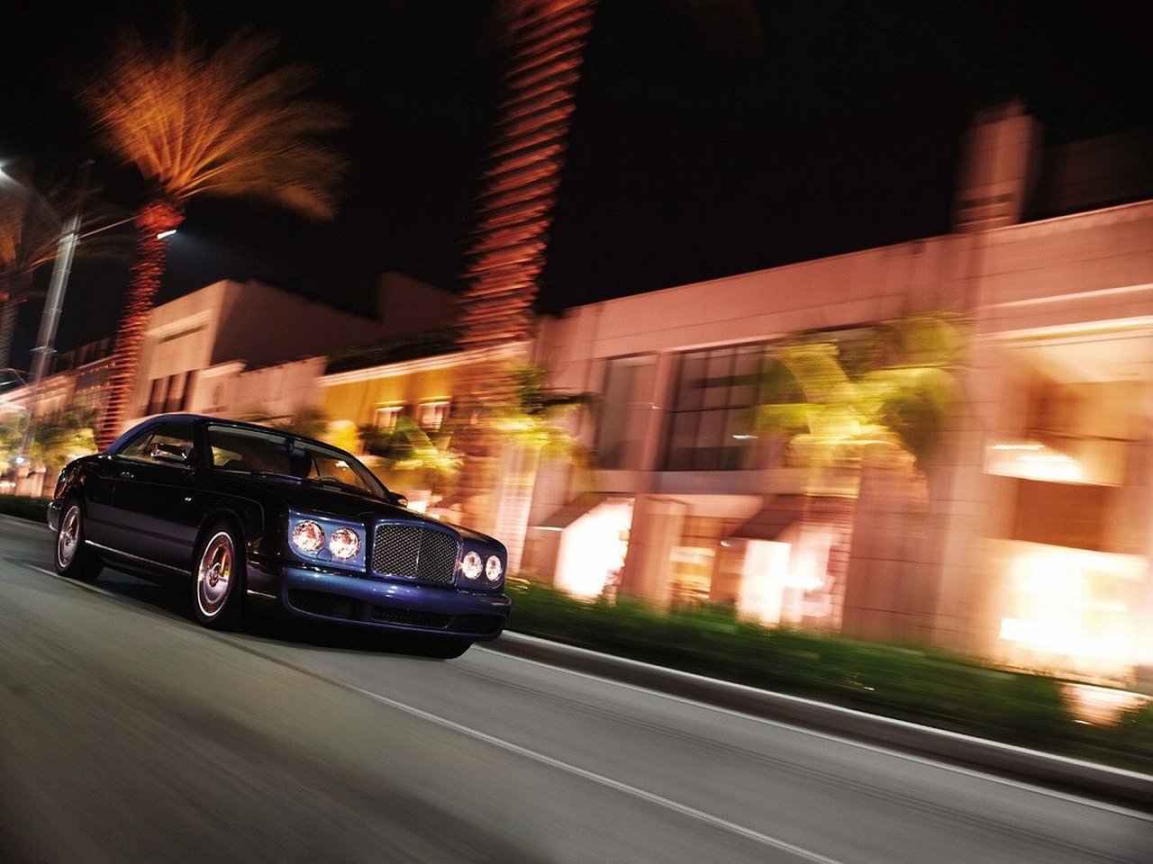 Black Bentley Azure driving past illuminated palm-lined buildings at night, motion blur conveying speed.
