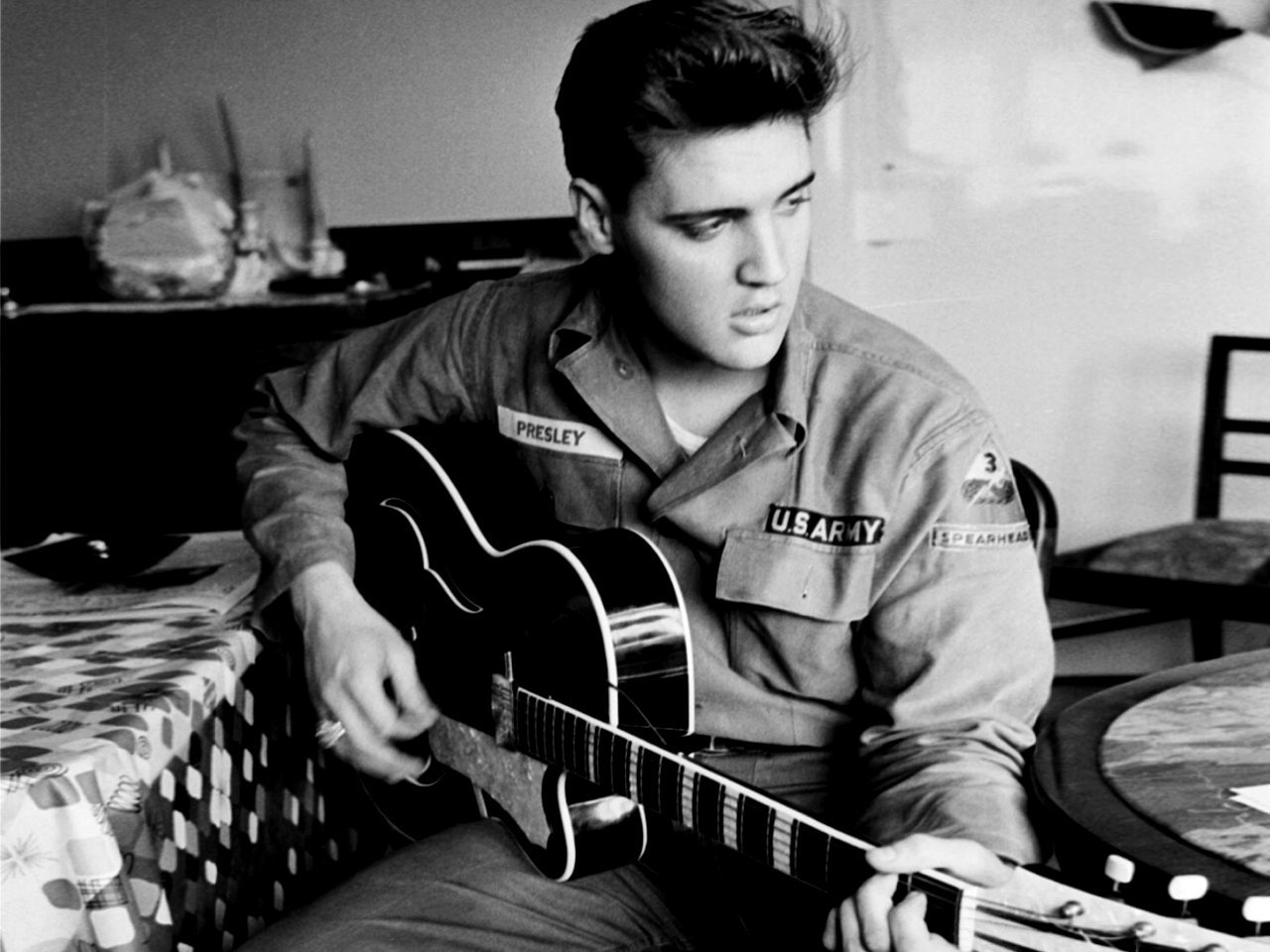 A black-and-white photo of Elvis Presley wearing a U.S. Army uniform, focused on playing his guitar while seated at a table, embodying the spirit of music and the 1950s era.