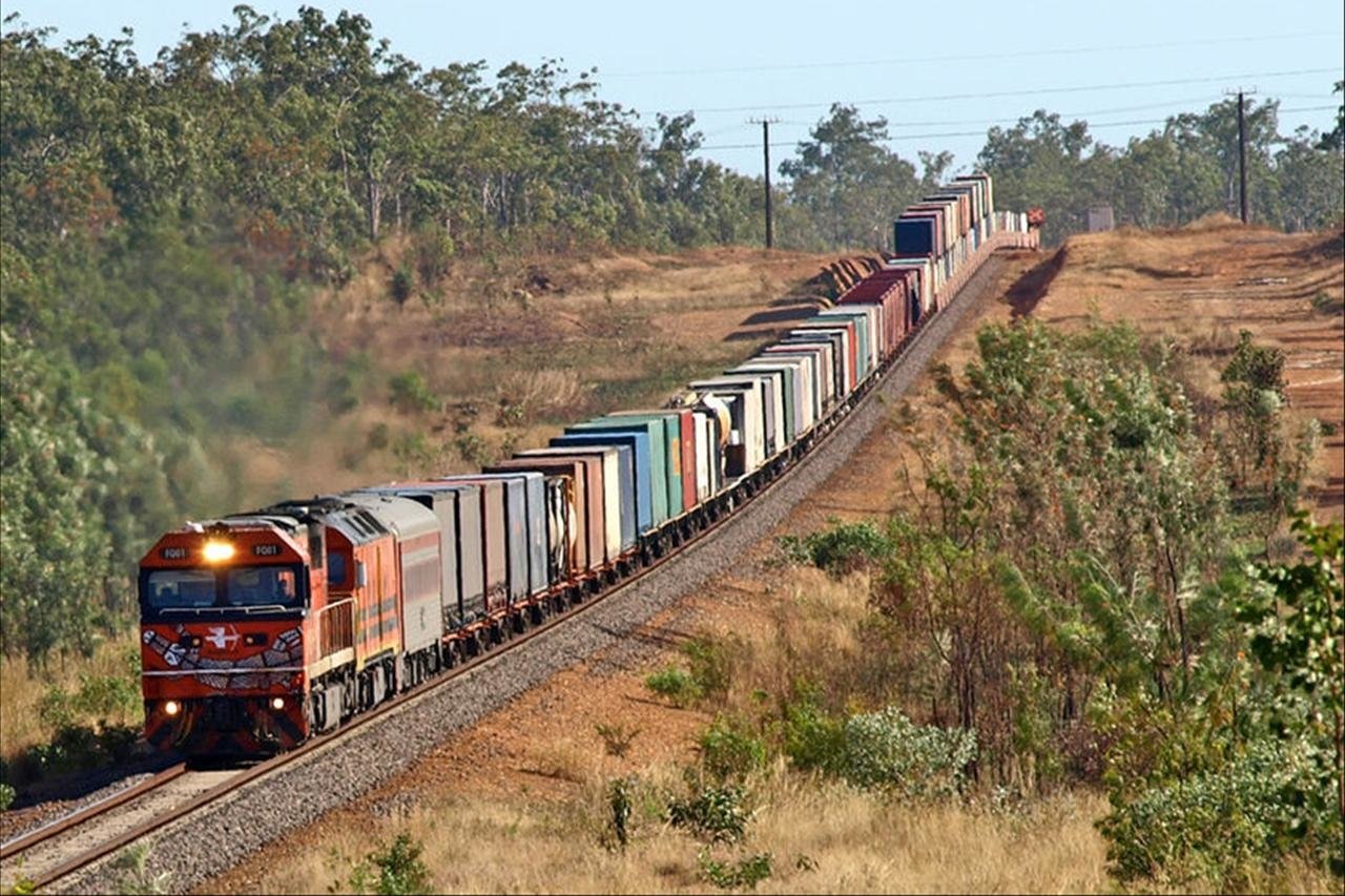 Long freight train vehicle hauling stacked containers along winding rural tracks through dry scrubland.