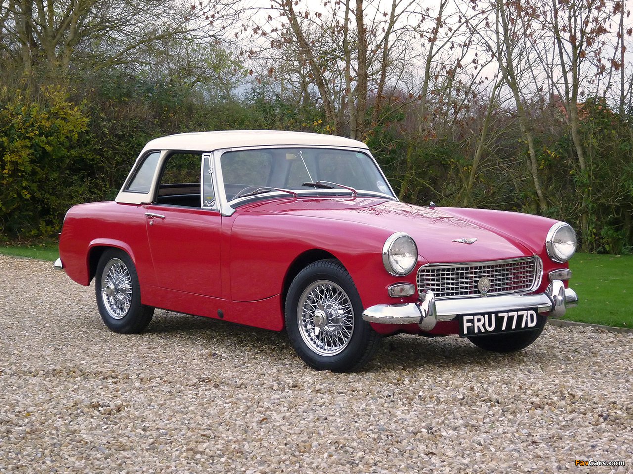 Classic red Austin-Healey Sprite sports car with white hardtop and chrome wire wheels, parked on gravel in front of trees.