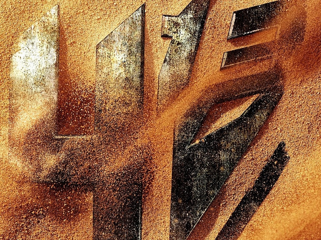 A close-up of the Transformers logo, partially veiled in sand, representing the movie Transformers: Age of Extinction. The metallic emblem stands out against the earthy background.