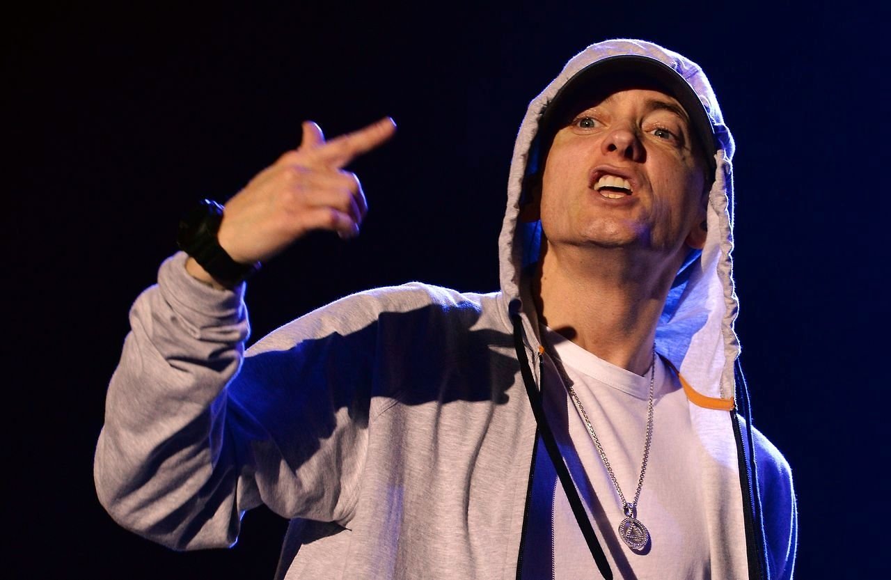 Eminem performs passionately on stage, wearing a hooded jacket and a pendant necklace, with intense expression and hand gesture emphasizing his music.