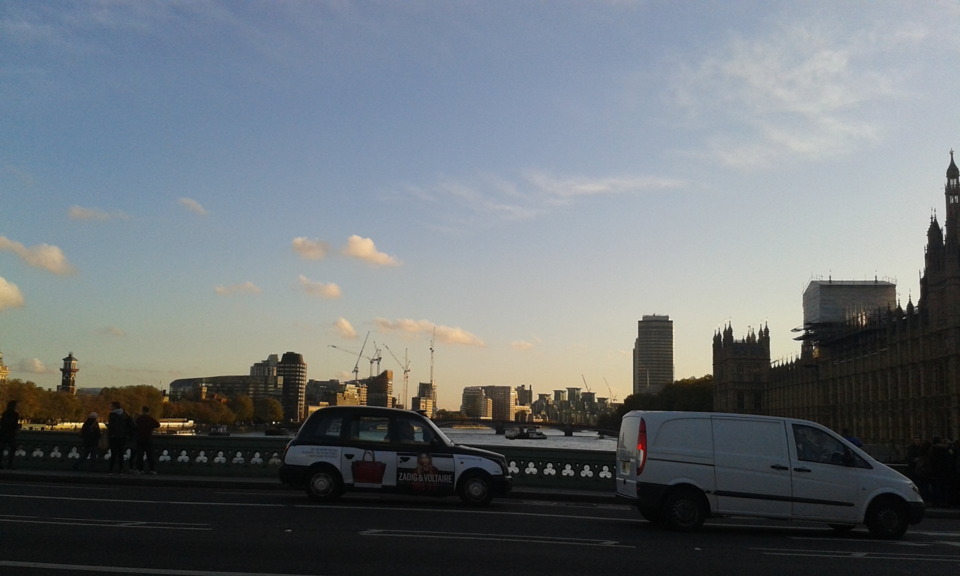A clear sky above a London street with a black taxi and a white van driving past historic buildings along the river.