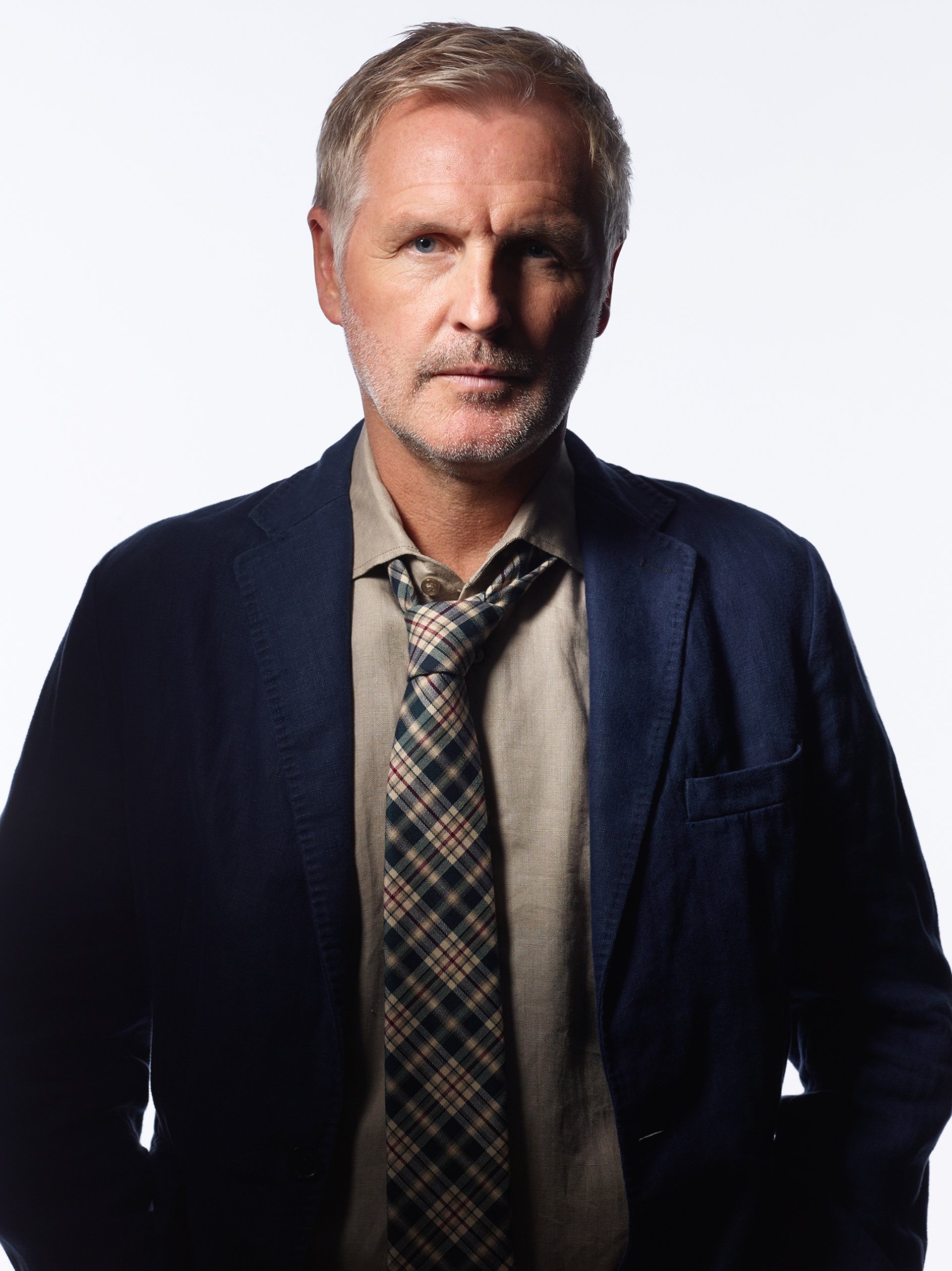 A confident Andrew Airlie poses in a stylish dark blazer and patterned tie, set against a light backdrop, showcasing his distinguished presence.