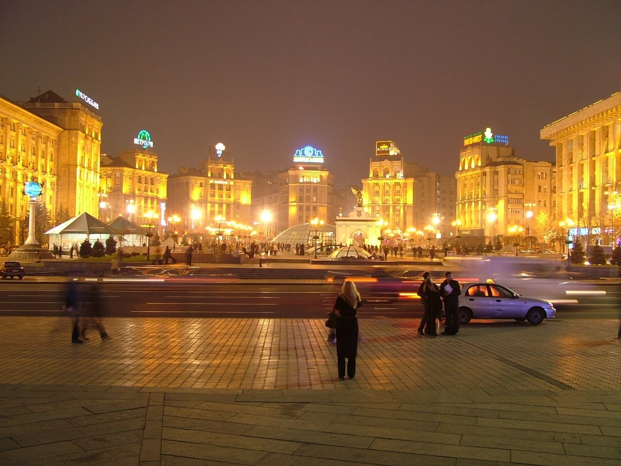 Night scene in Kiev: man-made illuminated buildings surrounding a busy paved plaza with pedestrians and cars.