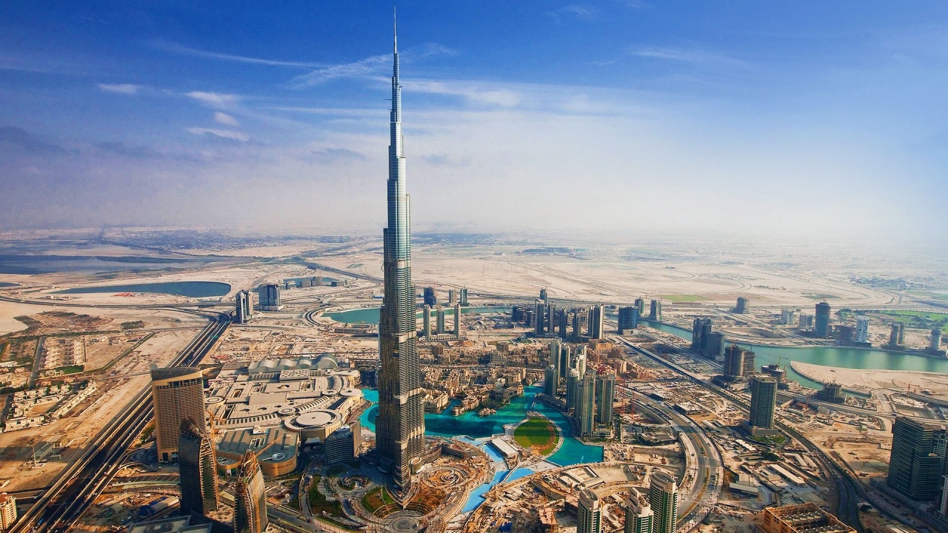 Aerial view of Dubai showcasing the iconic Burj Khalifa and surrounding man-made structures, set against a clear blue sky and desert landscape.