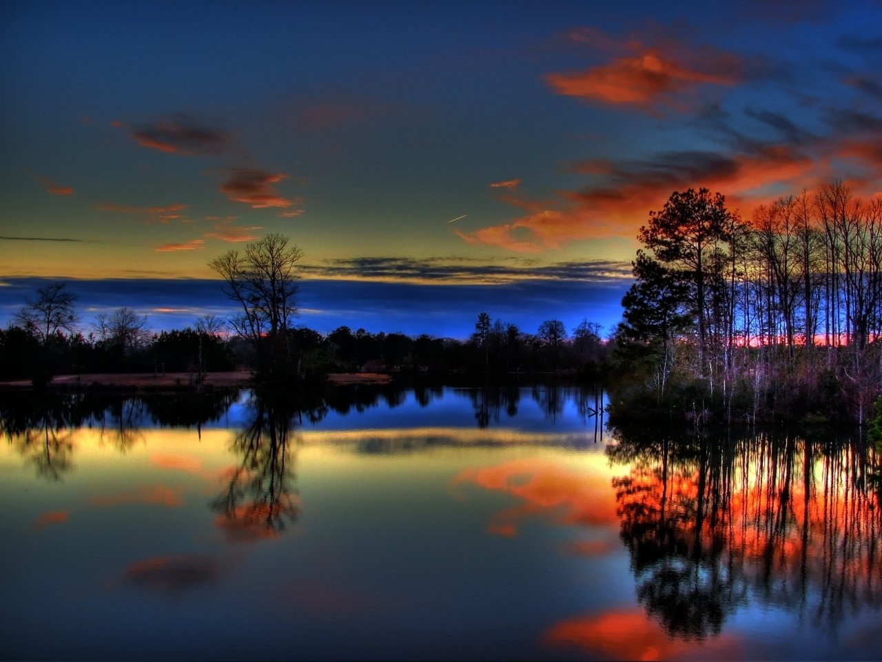 A serene landscape at dusk features vibrant clouds reflecting in calm waters, surrounded by silhouetted trees, creating a captivating nature scene.