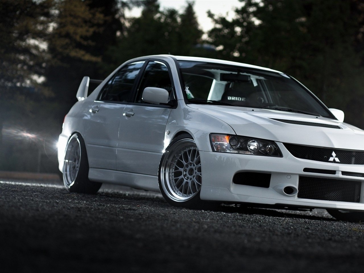A sleek white Mitsubishi Lancer Evolution parked on a dark surface, showcasing its sporty design and distinctive alloy wheels against a backdrop of trees.