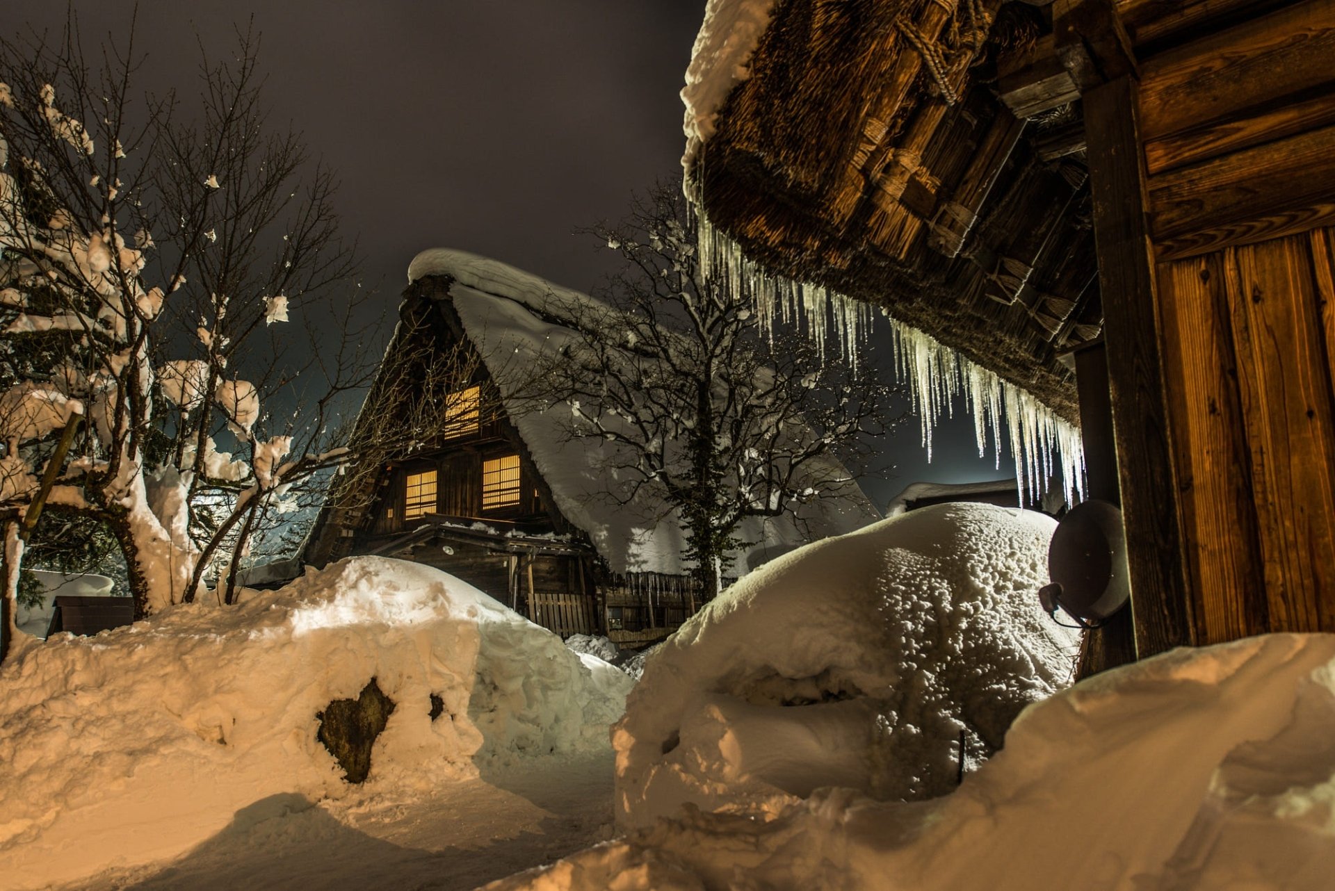 Snow-covered traditional Japanese houses with icicles hanging from the roof, softly illuminated by warm light on an evening in winter.