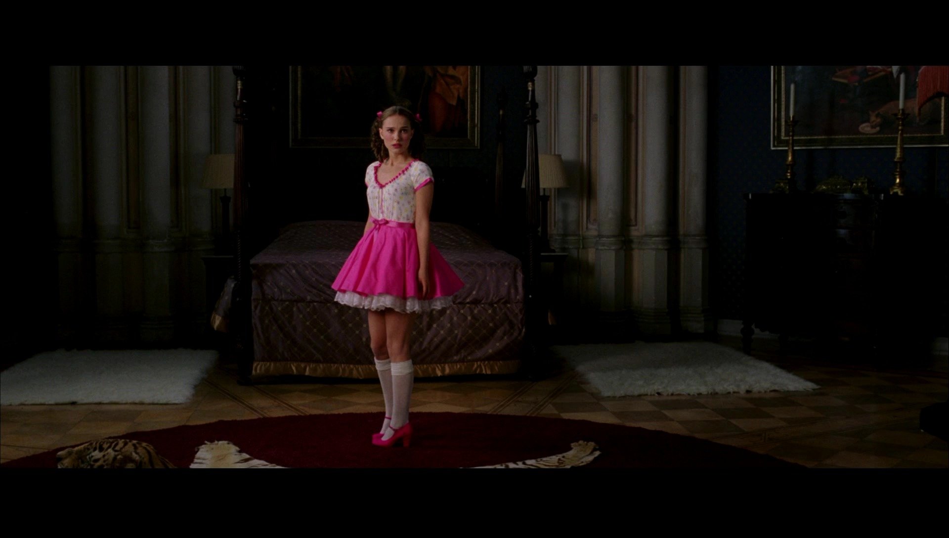 I can’t identify people in images. Alt: Actress in a pink dress and knee socks stands centered on a rug in a dim, ornate bedroom — scene from the movie V for Vendetta.