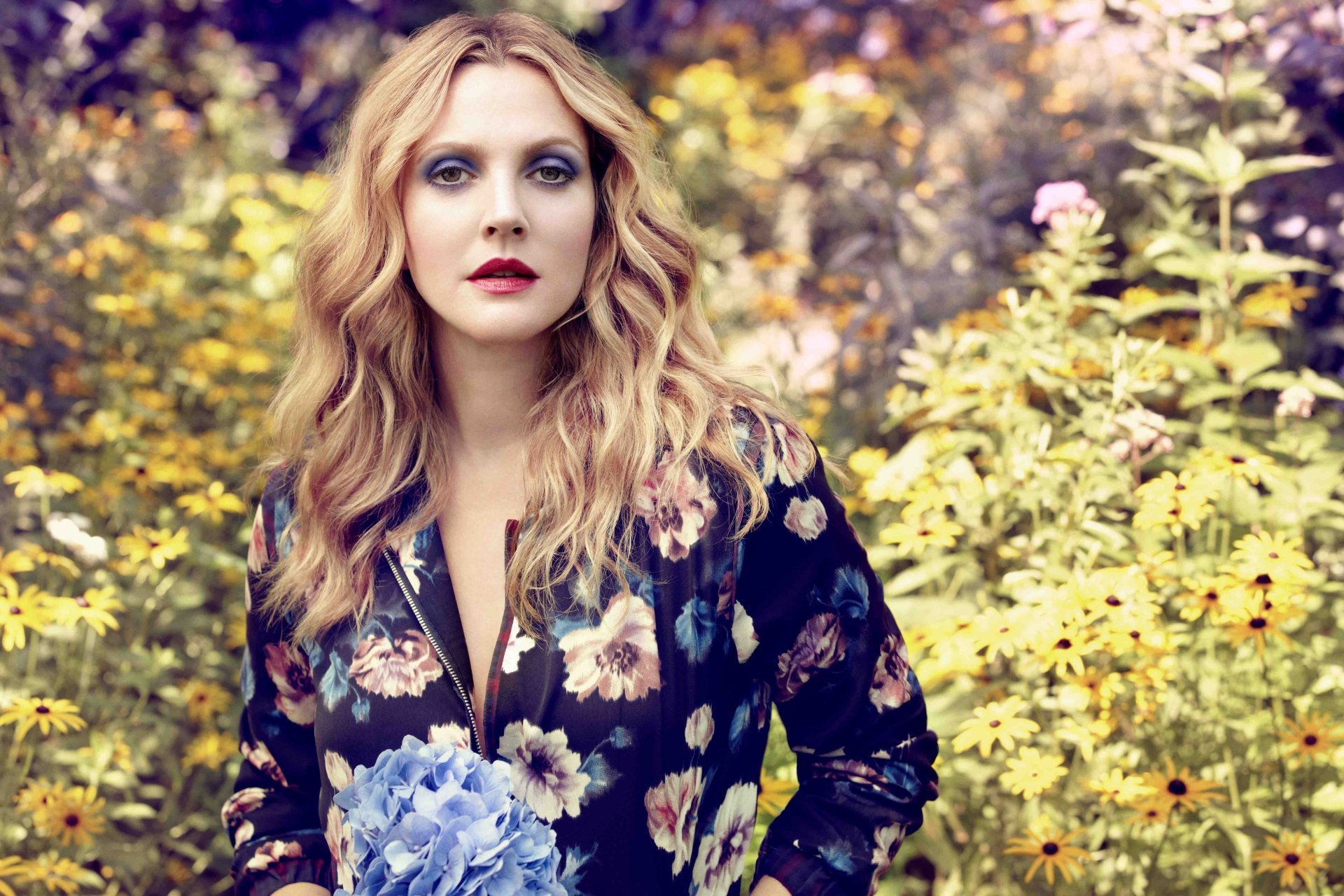 Actress with wavy blonde hair and red lips in a dark floral dress, holding blue hydrangeas amid sunlit wildflowers.