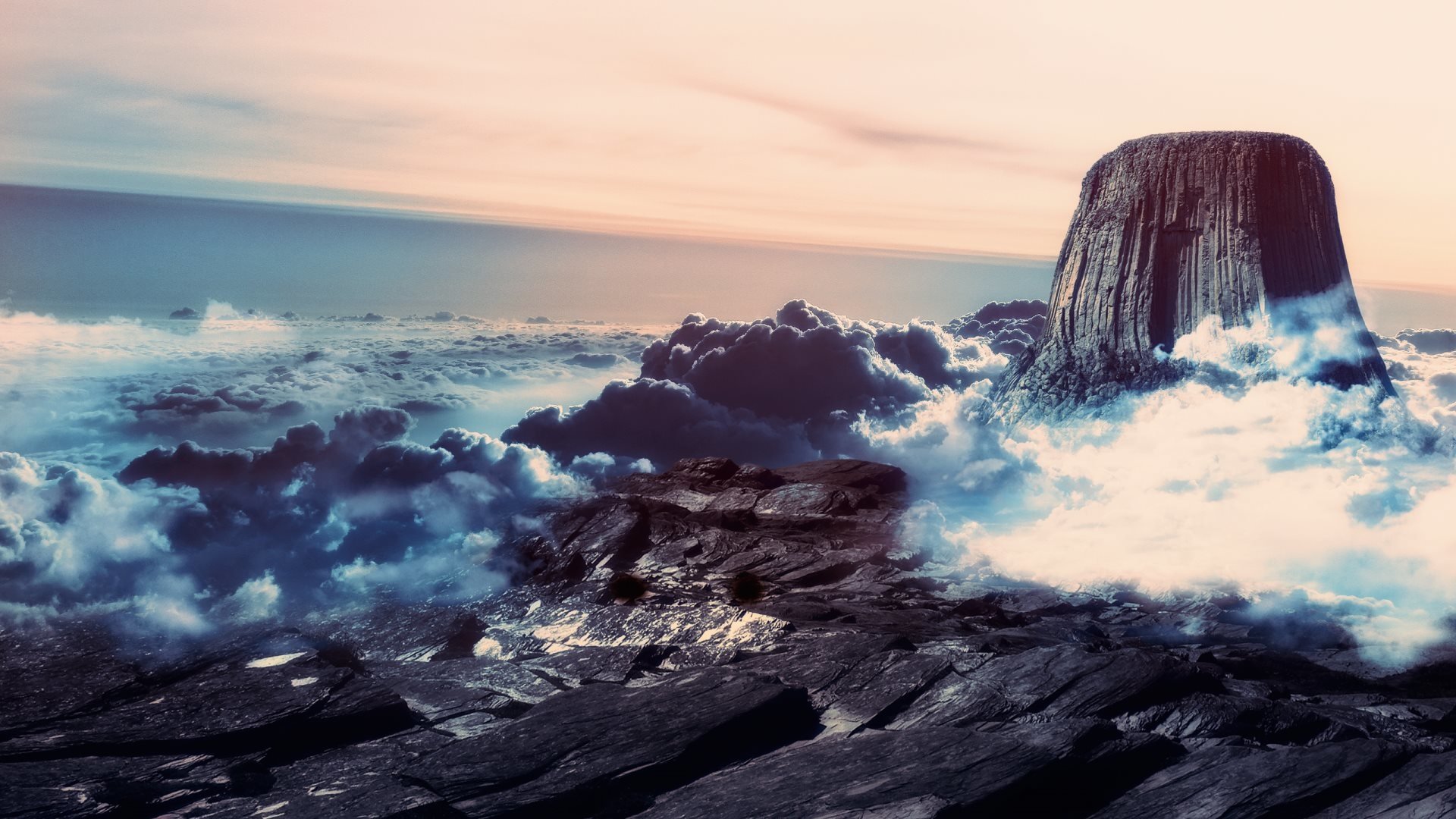 View of Devils Tower rising above rocky terrain surrounded by clouds under a soft, colorful sky at sunset.