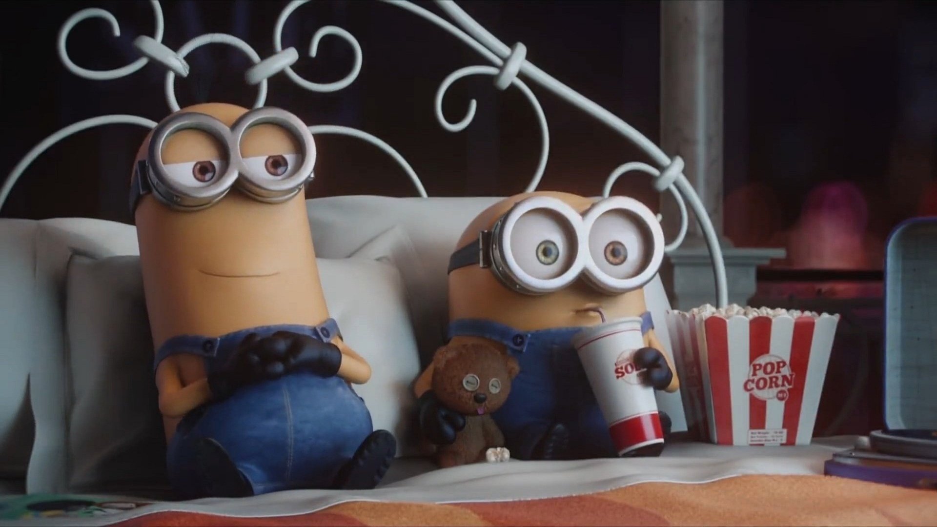 Movie scene showing two Minions sitting in bed—one holding popcorn and a drink, the other clutching a stuffed toy as they watch the film.