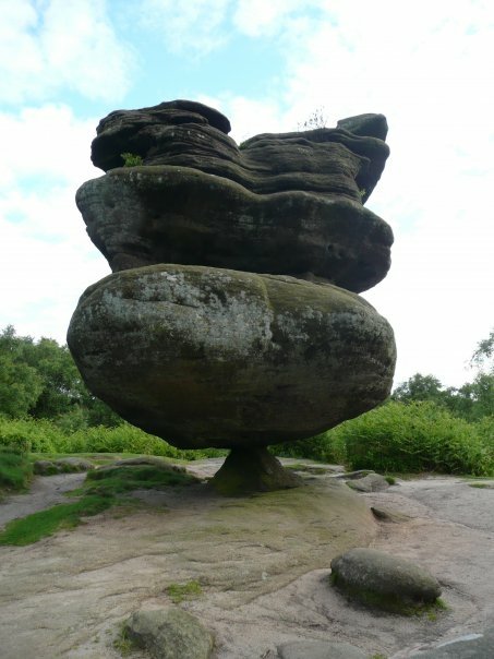 Mushroom-shaped balanced sandstone rock in Yorkshire nature, rising from moorland with green shrubs beneath a cloudy blue sky.