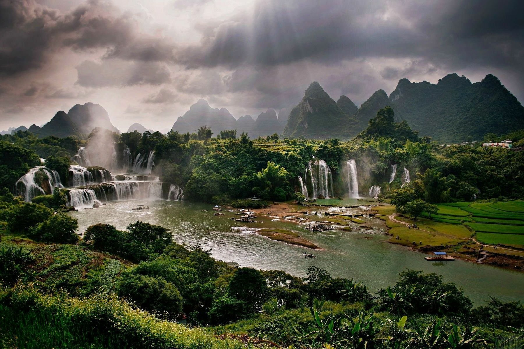 A breathtaking landscape showcasing cascading waterfalls amidst lush greenery and dramatic mountains, illuminated by beams of sunlight breaking through cloudy skies.