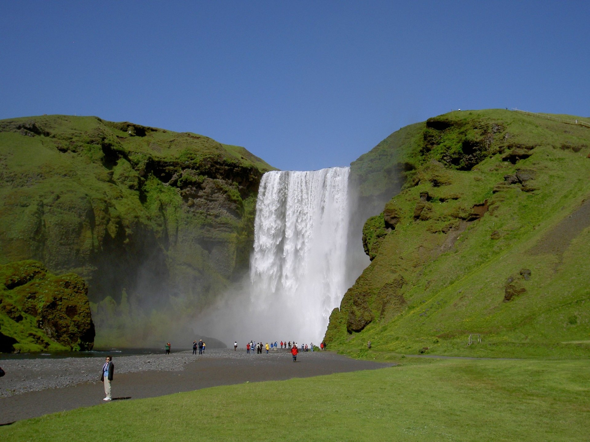 A powerful waterfall cascades between lush green hills under a clear blue sky, with people gathered near the water’s edge in this serene nature scene.
