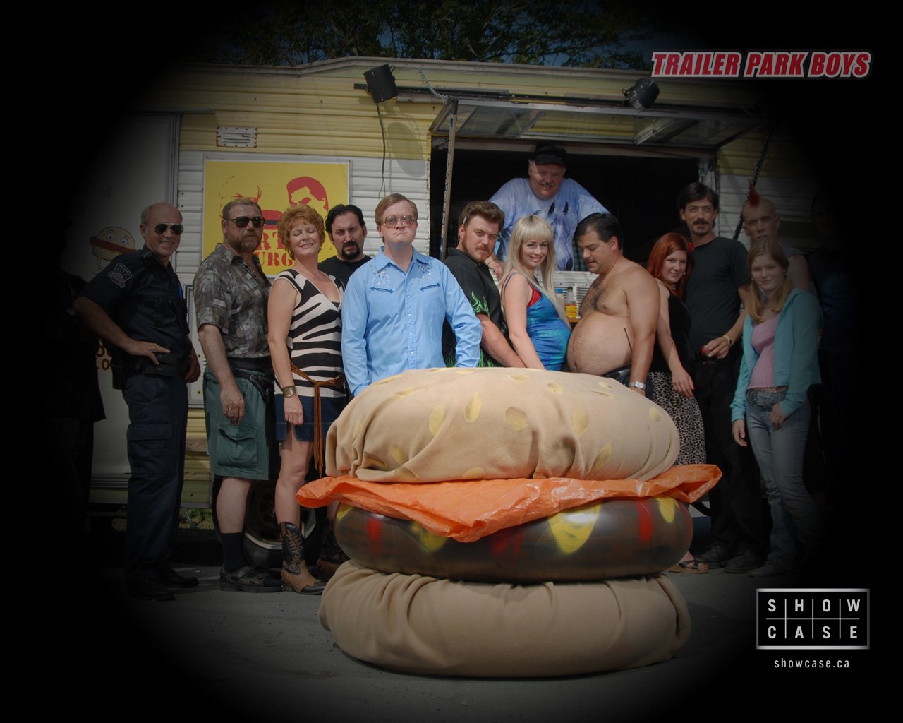 A group of characters from the TV show Trailer Park Boys poses playfully in front of a trailer, with a large, humorous burger prop in the foreground.
