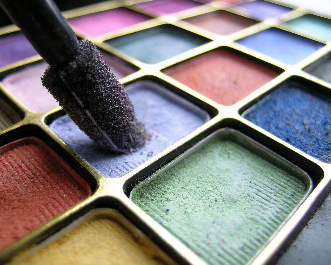 A close-up of a makeup applicator touching a vibrant purple shade, surrounded by an array of artistic colors in various squares, showcasing texture and creativity.