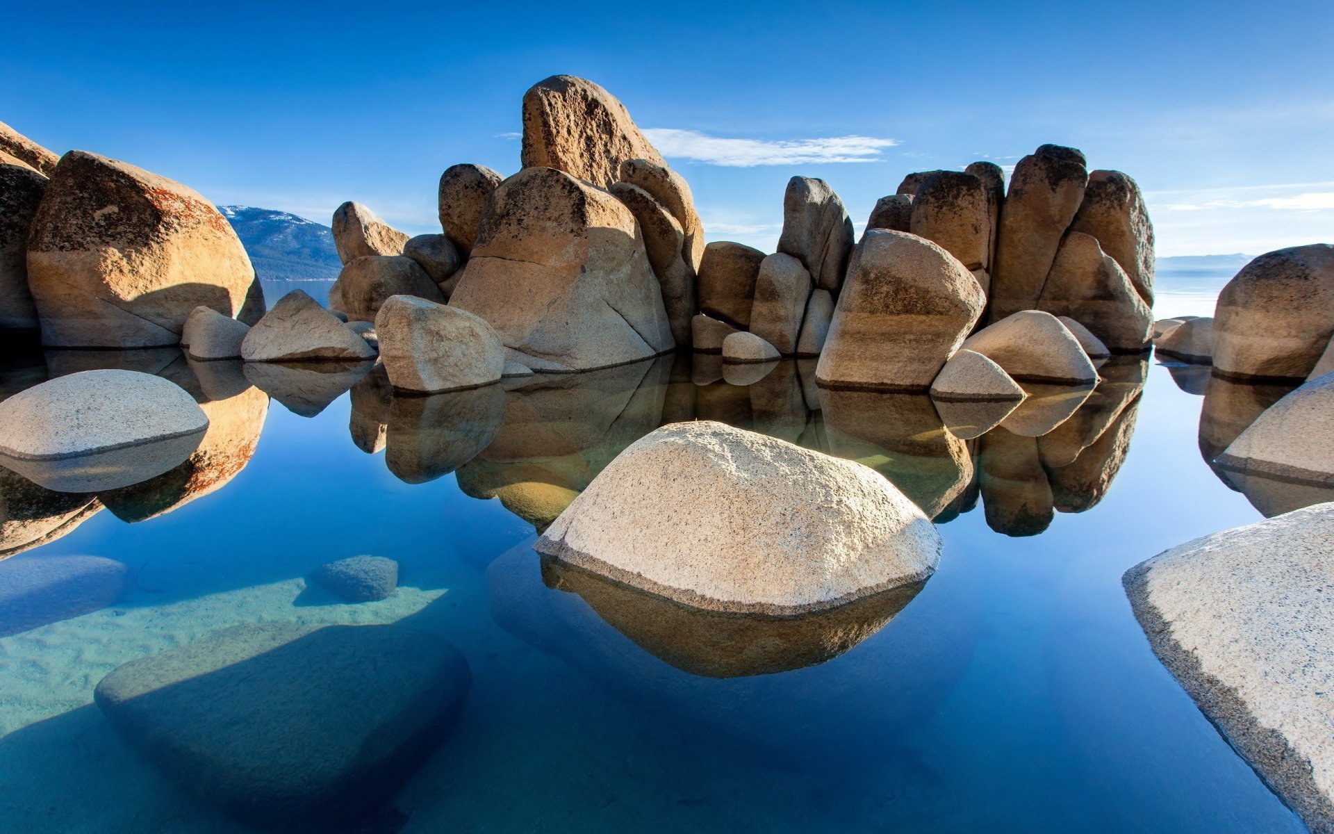 A serene natural scene featuring large rocks reflected in the calm, clear water under a bright blue sky.