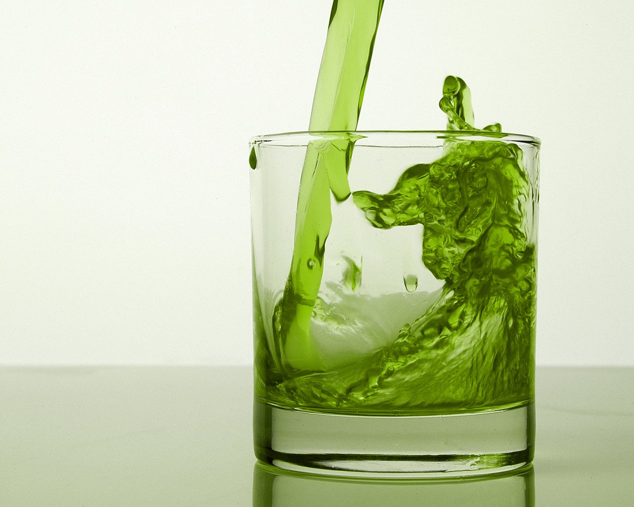 A vibrant green liquid is artistically pouring into a glass, creating dynamic waves and splashes, showcasing an elemental and refreshing aesthetic.