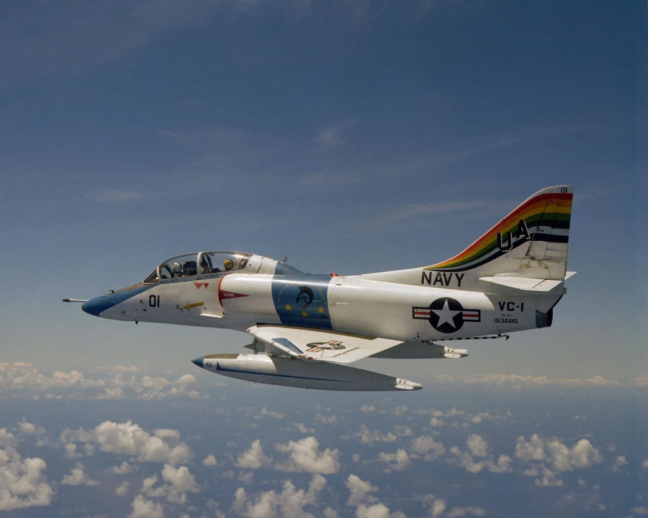 A Douglas A-4 Skyhawk jet, showcasing a colorful tail design, soars through a clear blue sky with fluffy white clouds in the background, representing military aviation excellence.