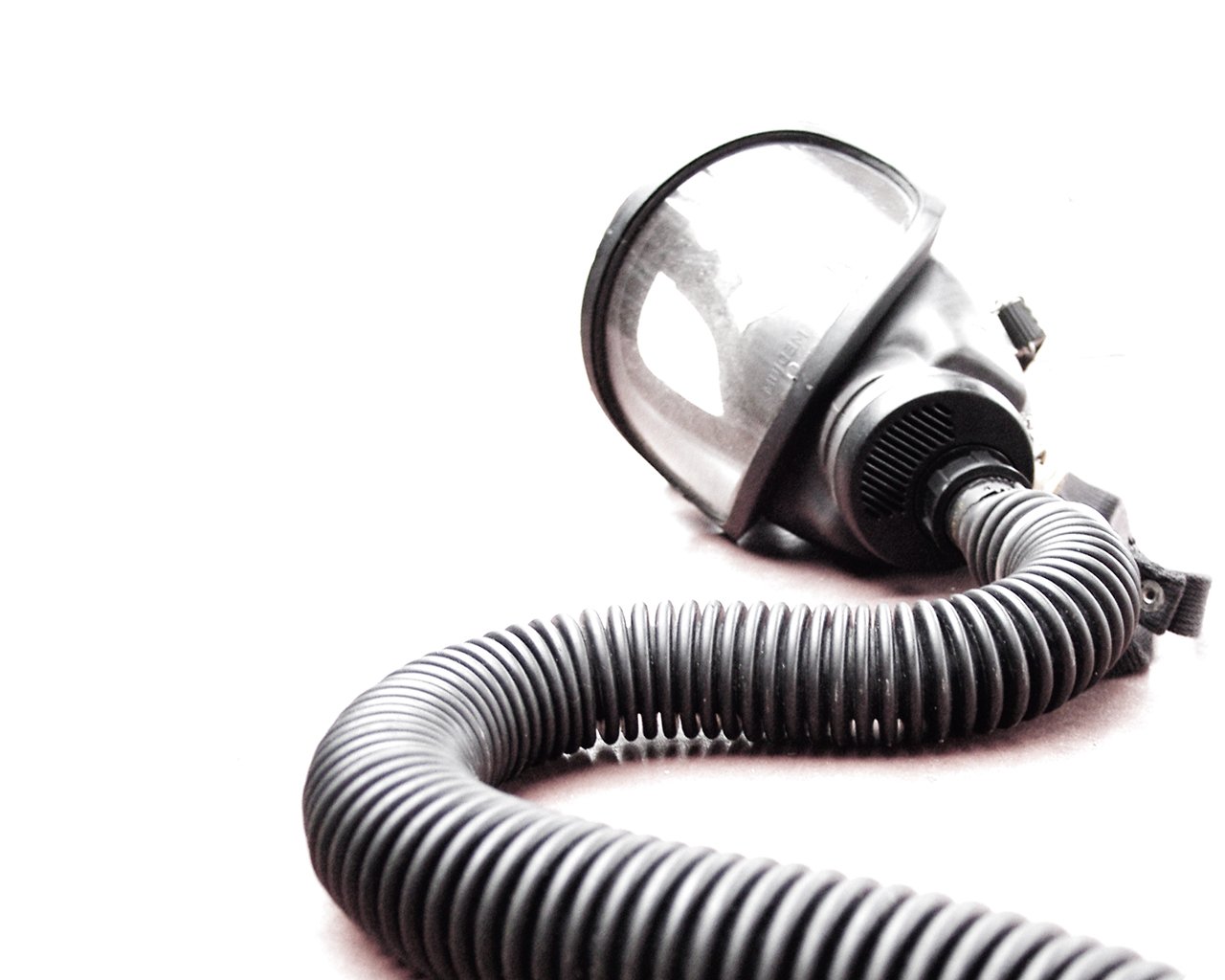 A military gas mask with a flexible hose lies against a light background, emphasizing its design and functionality for protection in hazardous environments.