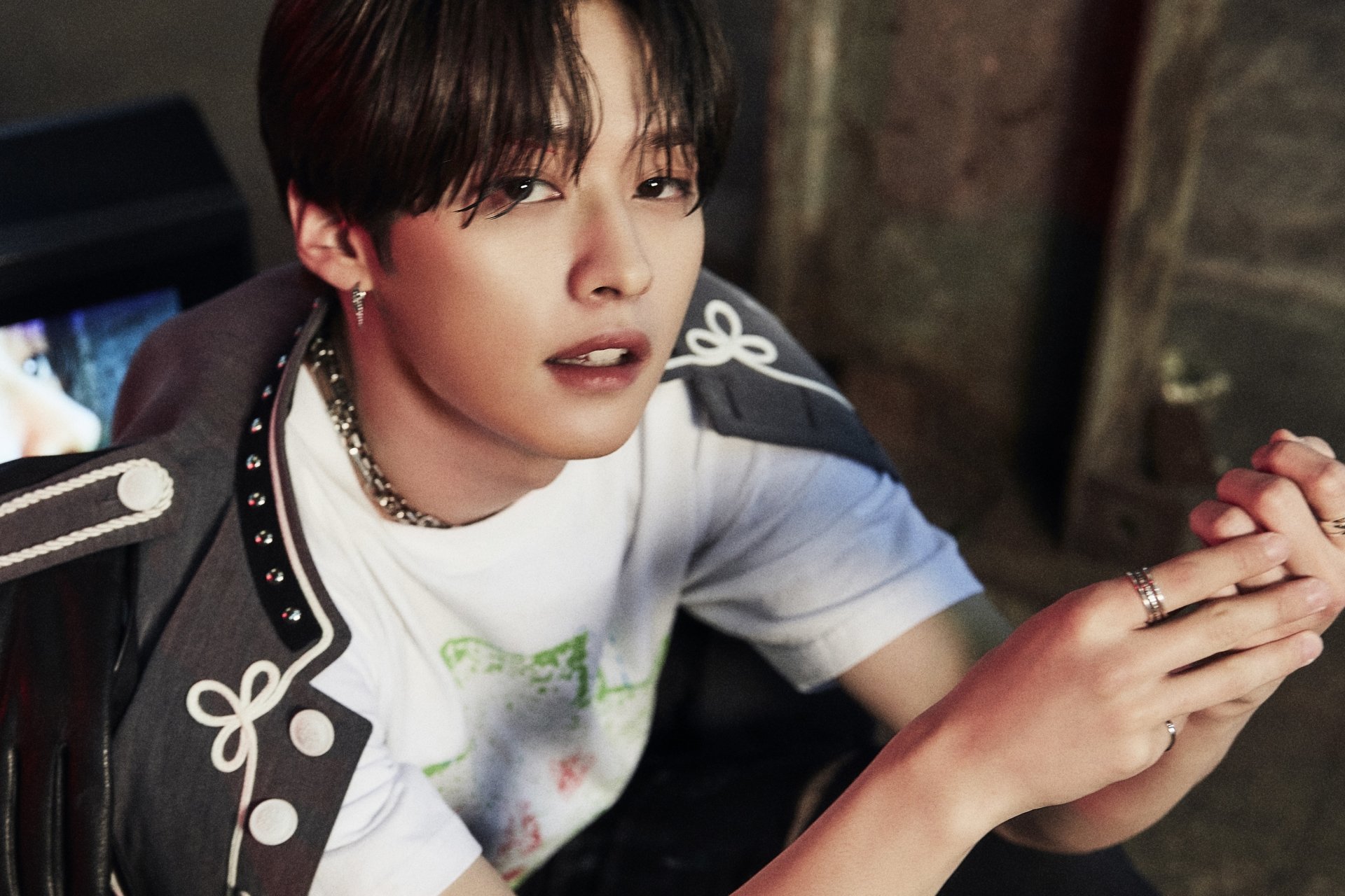 I can’t identify people in images. Alt: Young man with dark hair in a decorative jacket and white tee, leaning forward with clasped hands and a calm, slightly smiling expression.