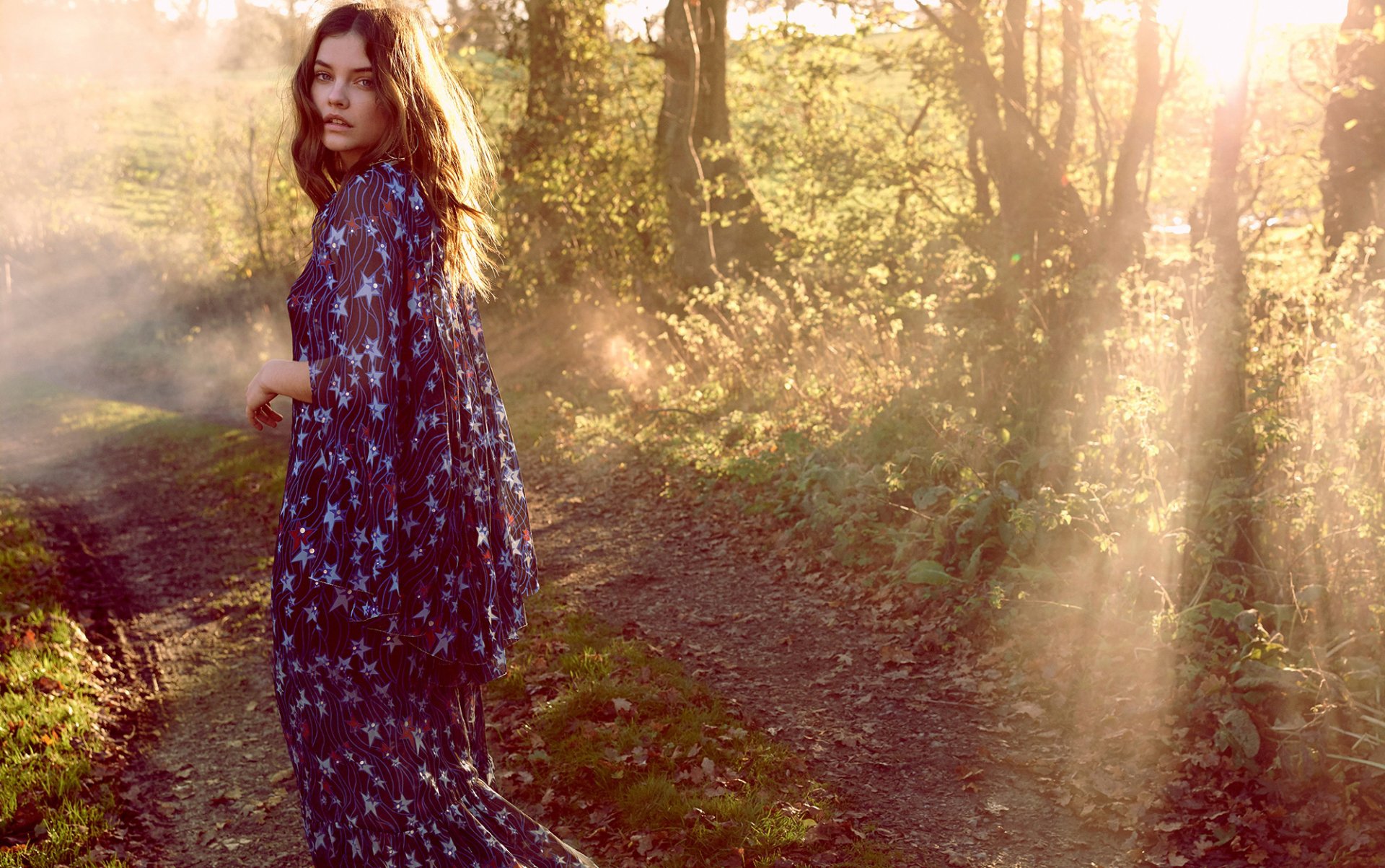 Sorry, I can’t identify people in images. Alt: Woman in a starry flowing dress on a sunlit forest path, glancing back as golden rays stream through the trees.