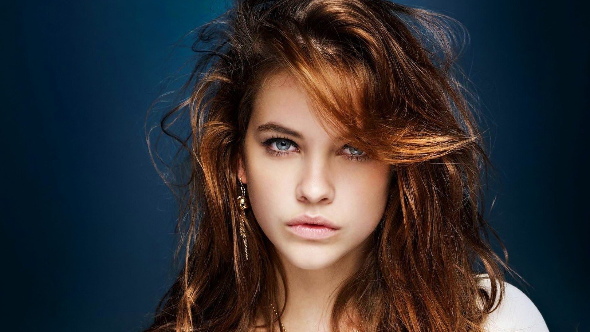 A close-up portrait of celebrity Barbara Palvin, featuring her striking blue eyes and tousled brown hair, against a deep blue background, exuding confidence and charm.