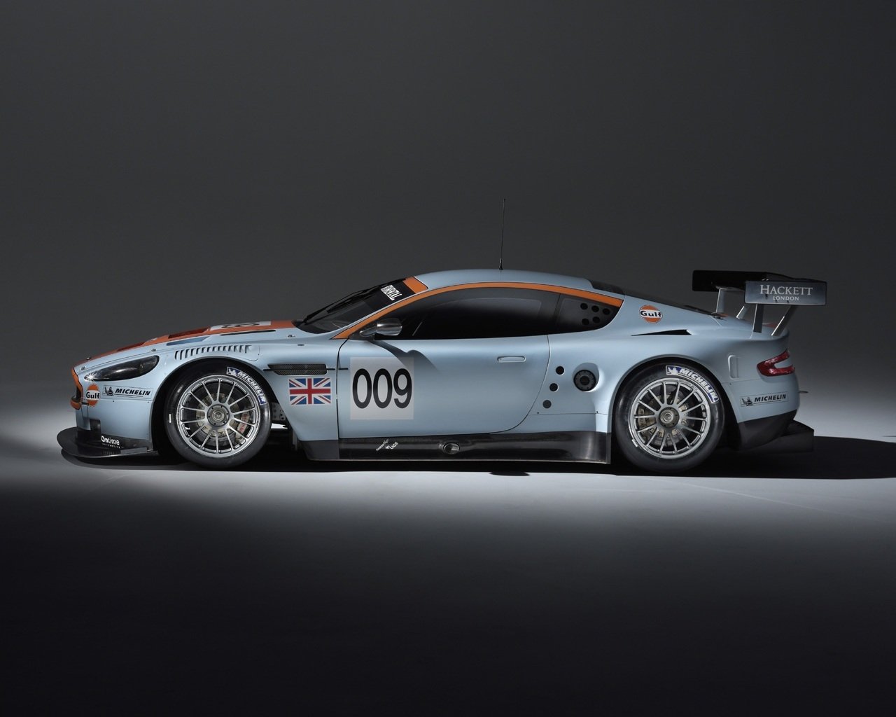 Side view of a silver Aston Martin DBR9 race car with the number 009 and British flag decals, showcased against a dark background.