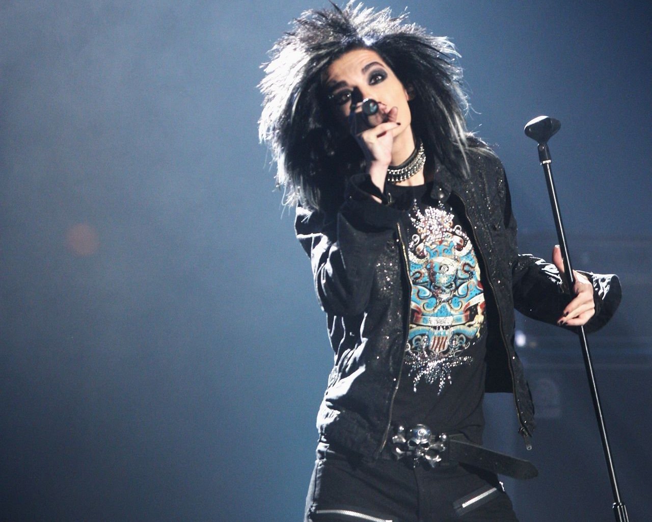 Lead singer of Tokio Hotel performing on stage with dramatic lighting, holding a microphone and wearing rock-style clothing and accessories.