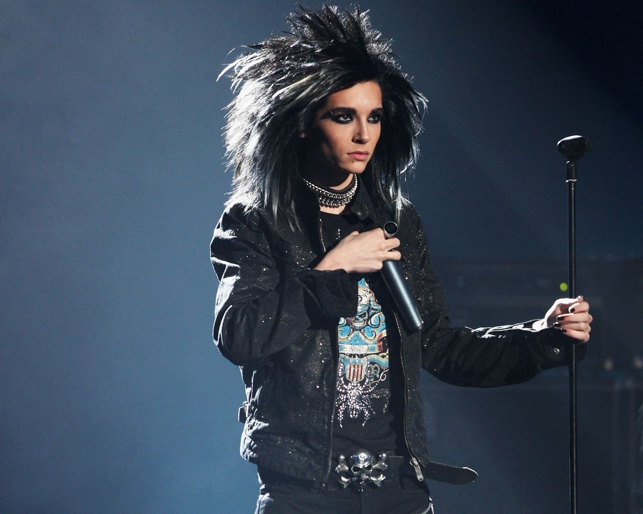 A musician from Tokio Hotel performs on stage, showcasing a dramatic hairstyle and dark fashion, holding a microphone amid dramatic lighting.
