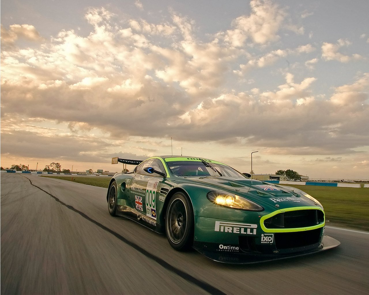 Aston Martin DBR9 racing on a track under a dramatic sky, showcasing its sleek design and vibrant colors as it speeds forward.