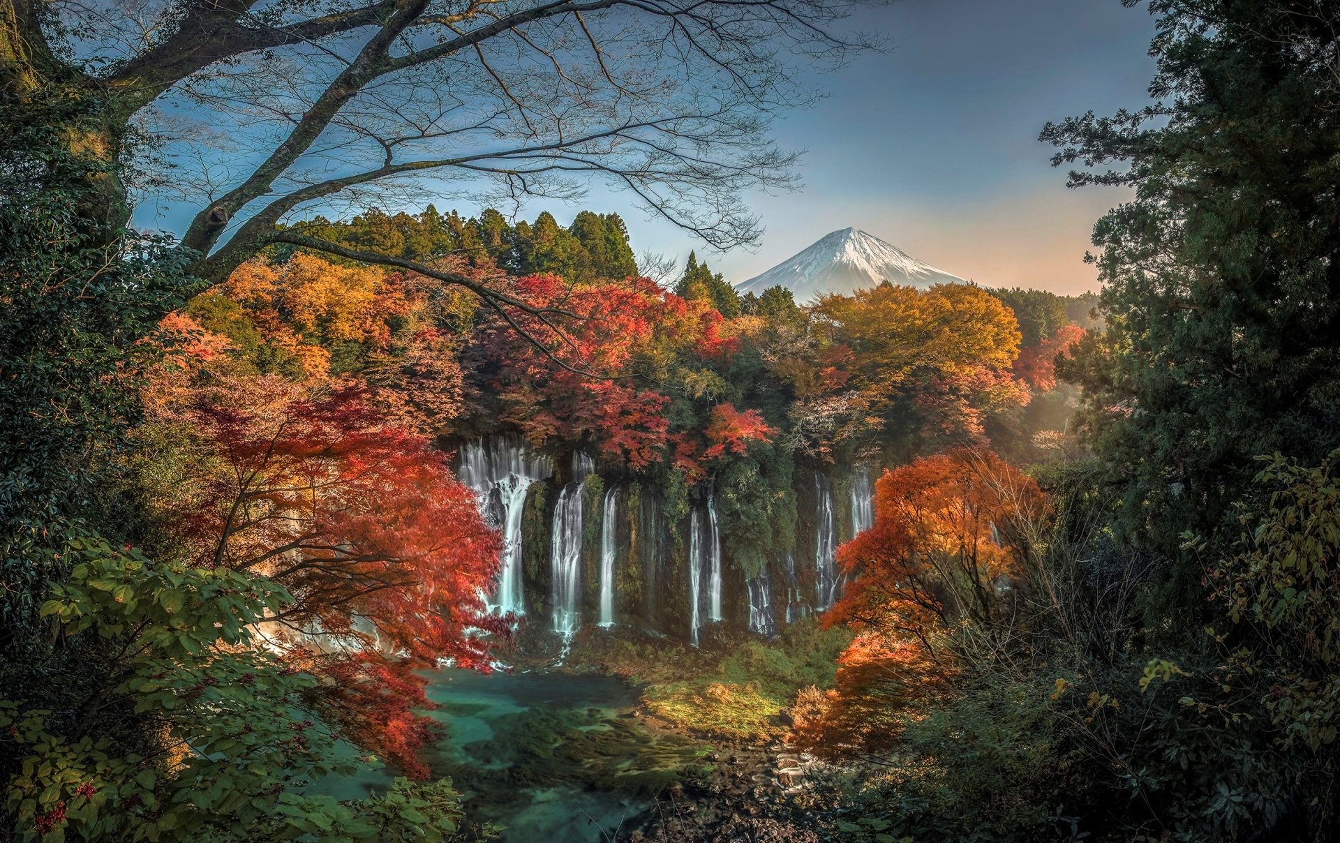 A vibrant autumn scene in Japan, featuring colorful foliage around a serene lake, with Mount Fuji rising majestically in the background and a waterfall cascading nearby.