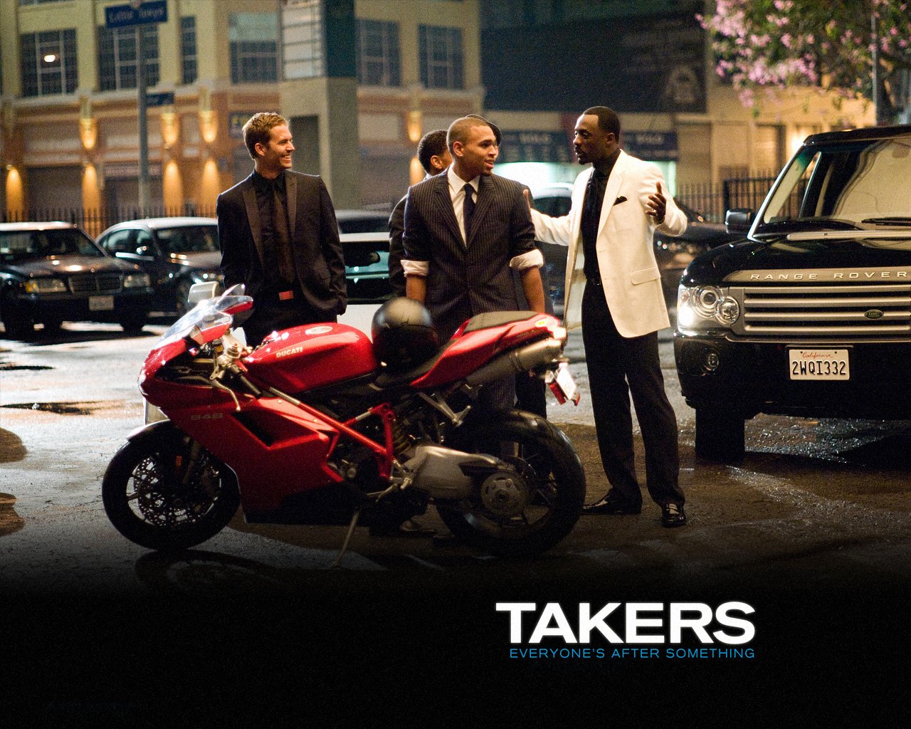 Three men in suits stand near a red motorcycle on a city street at night in a scene from the movie Takers.