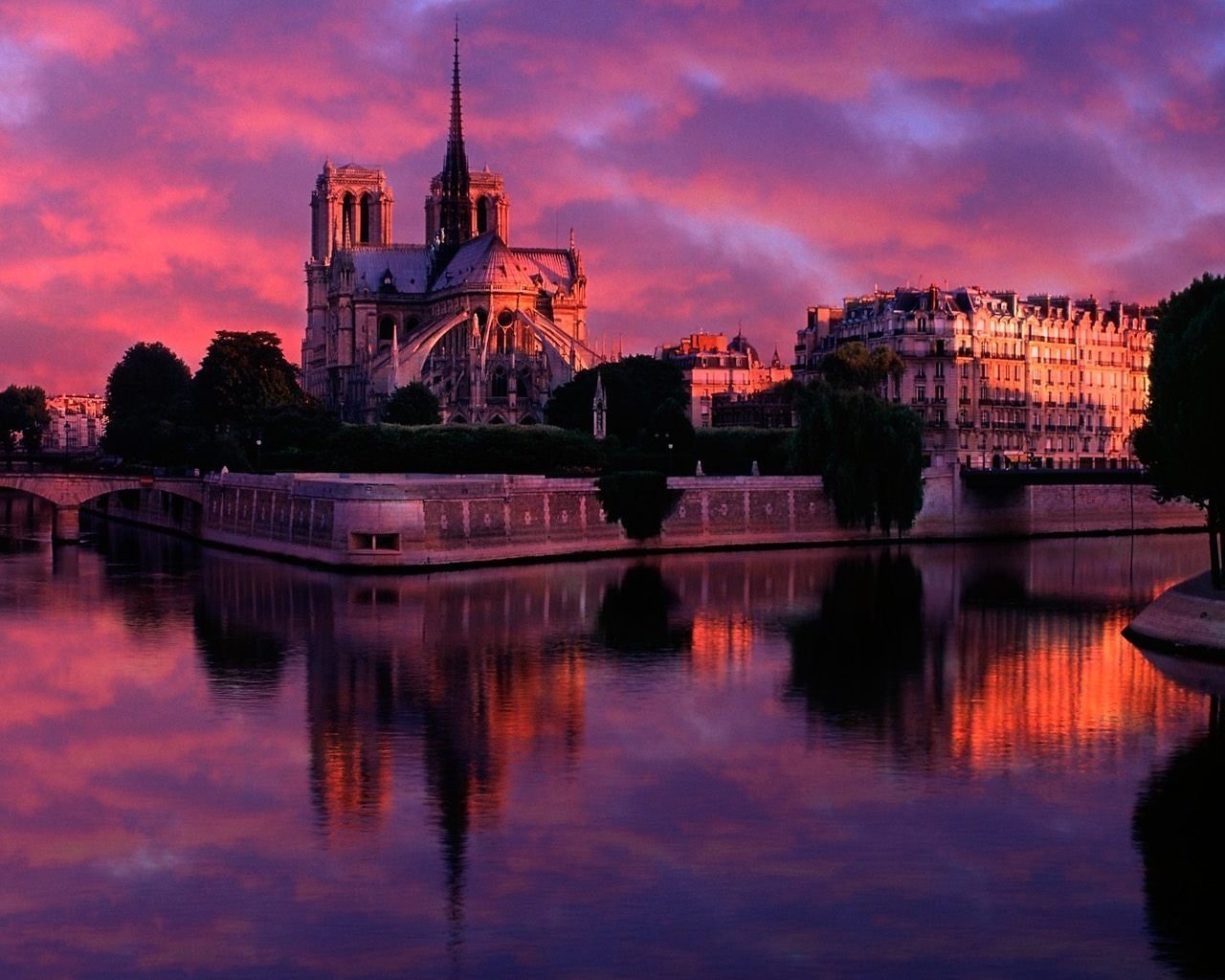 Notre-Dame de Paris illuminated at sunset, reflecting on the Seine River with a vibrant pink and purple sky.