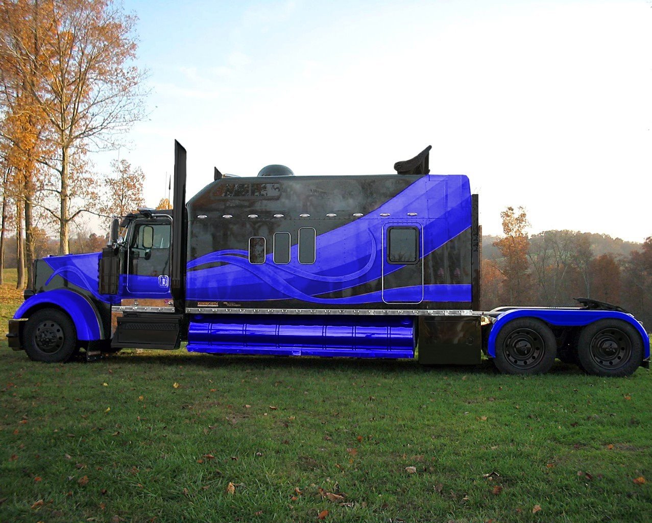 A striking Kenworth vehicle displays a bold blue and black design, parked on grassy terrain with autumn trees in the background, showcasing its powerful presence.