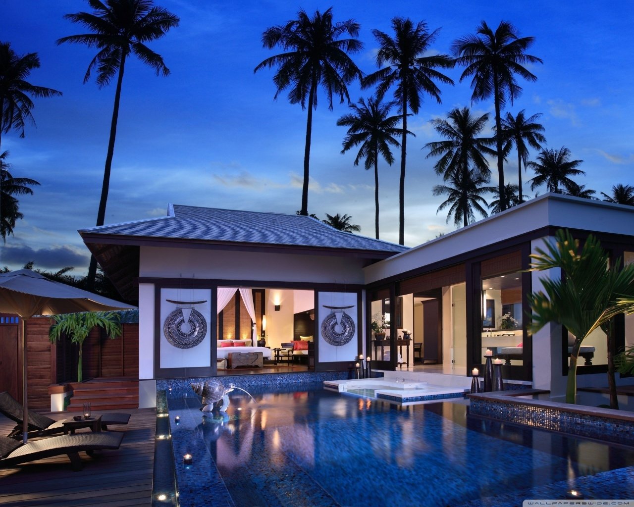 Modern villa at dusk with a man-made pool in the courtyard, open glass-front living areas, lounge chairs and tall palm trees against a deep blue sky.