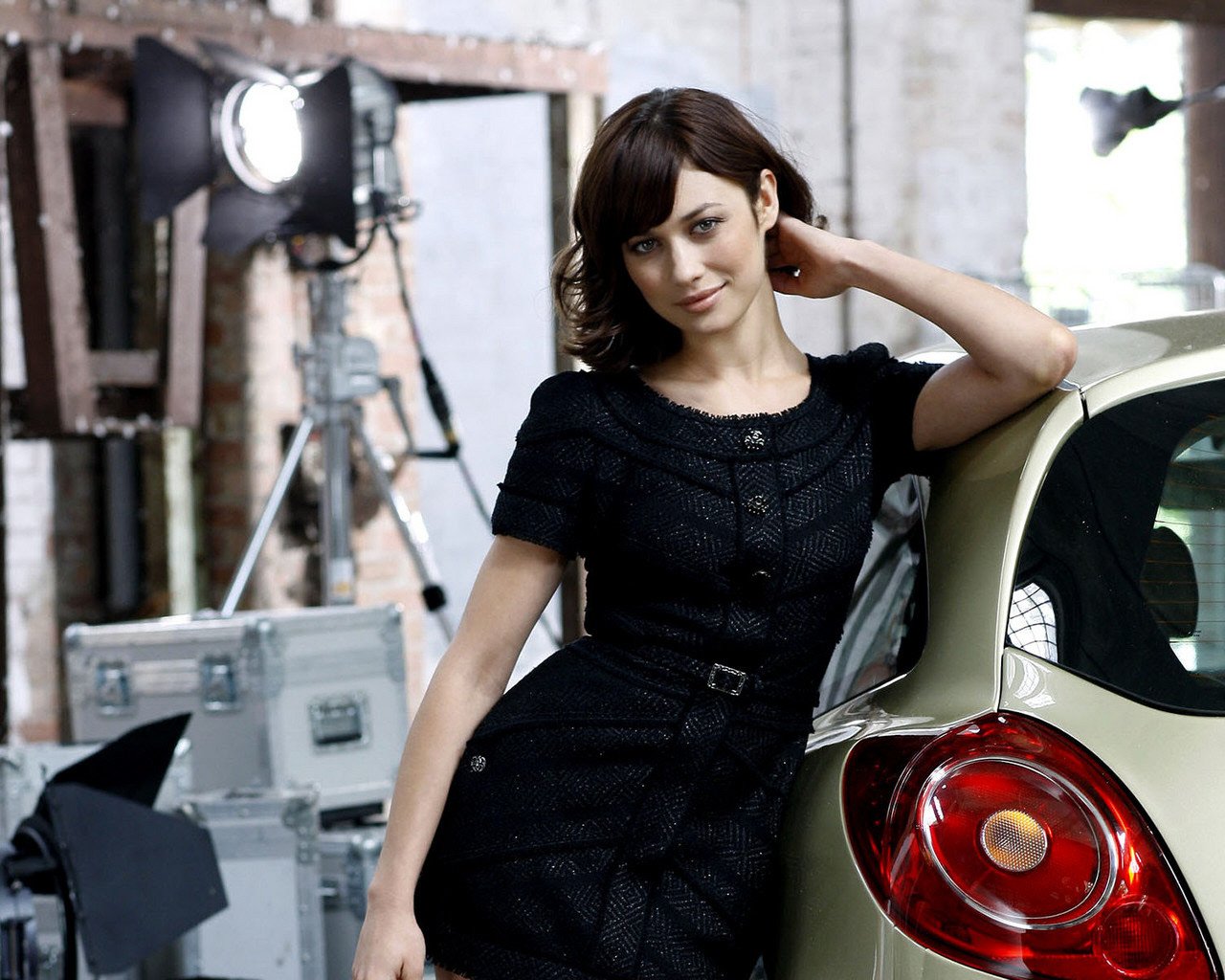 Celebrity Olga Kurylenko poses confidently in a black dress, leaning against a car in a stylish, industrial setting with studio lighting in the background.