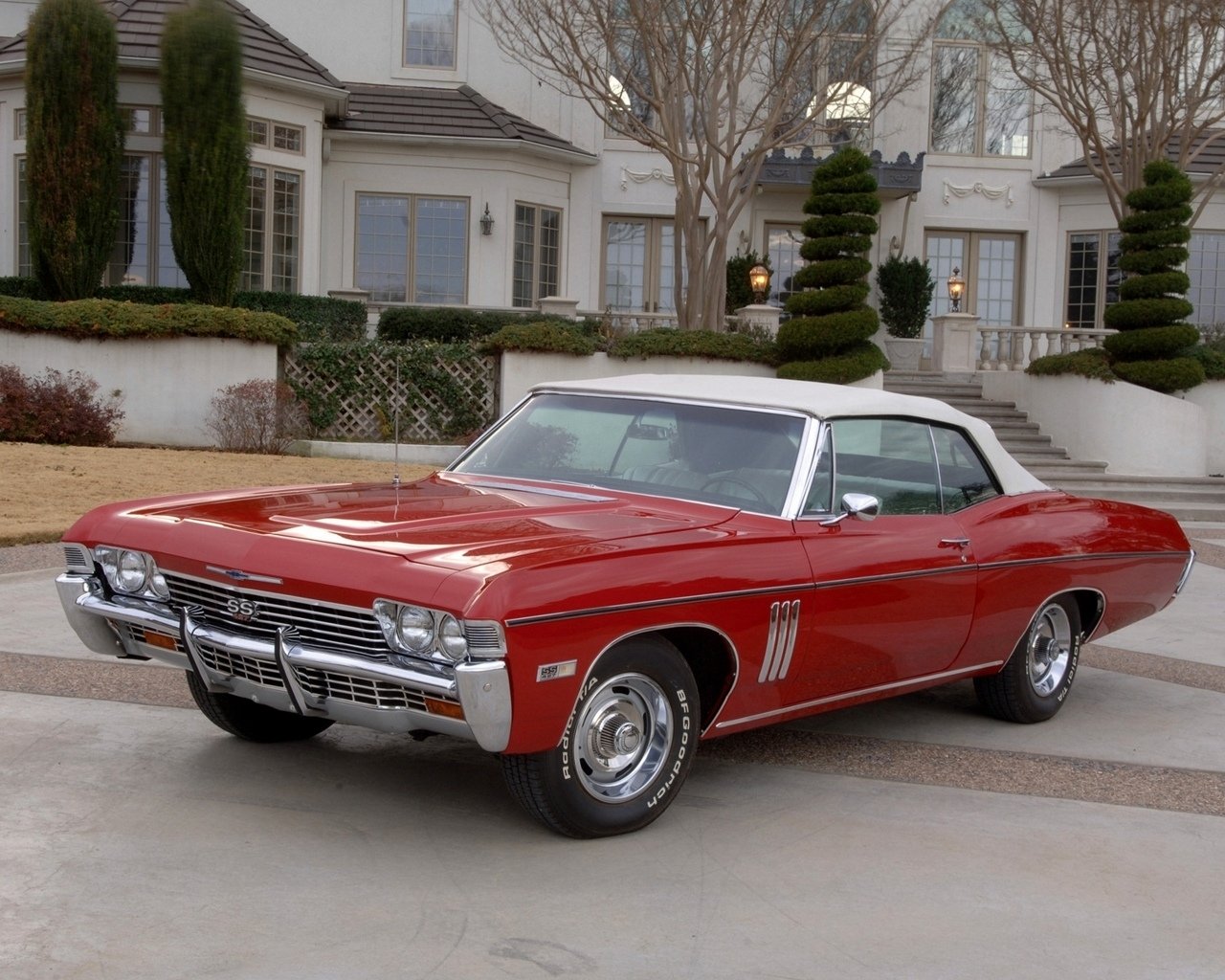A shiny red classic vehicle with a white convertible top is parked in front of a large, elegant house with manicured bushes.